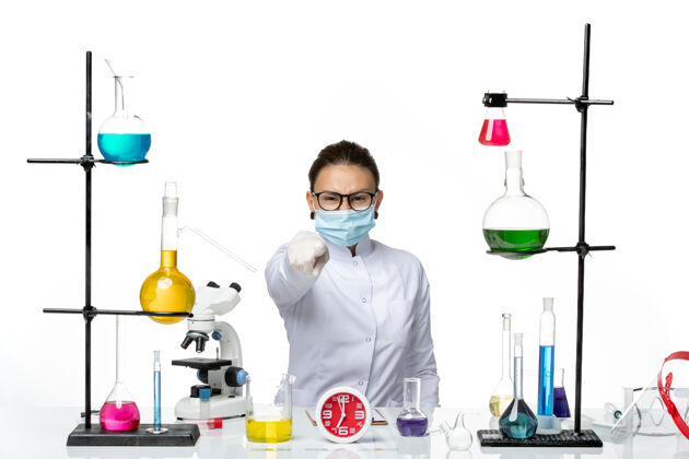 医学前视图：穿着医疗服的女化学家戴着面罩 坐在浅白背景上的溶液化学实验室病毒-喷溅飞溅前面实验室