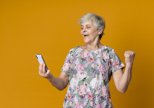 拳头快乐的老妇人举起拳头 看着隔离在橙色墙上的电话快乐老人举起来