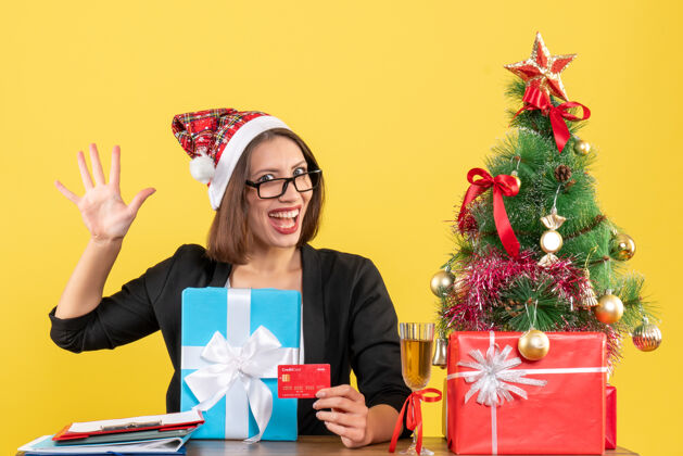 圣诞老人微笑迷人的女士穿着西装 戴着圣诞老人的帽子 戴着五副眼镜 手里拿着礼物和银行卡 在黄色的办公室里与世隔绝肖像帽子圣诞老人