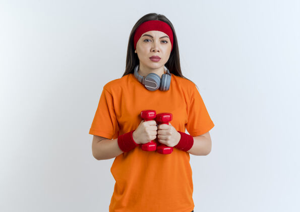 头带自信的年轻运动女性戴着头带和腕带 脖子上戴着耳机 看起来孤立地举着哑铃耳机年轻颈部