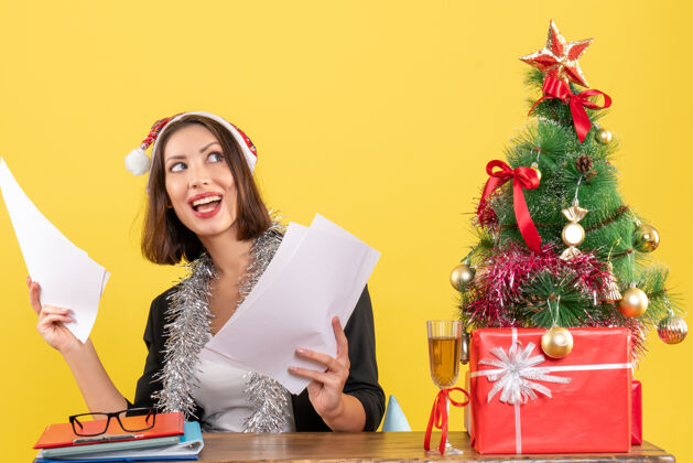 树心满意足 情绪集中的商务女士 穿着西装 戴着圣诞老人的帽子和新年装饰品 手里拿着文件 坐在办公桌旁 桌上放着圣诞树桌子满足年份