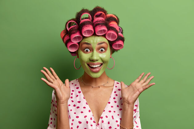 清洁非常高兴的女性模特儿举手掌做早操 用保湿绿色面膜复壮 戴卷发器 隔离绿色 随意穿戴过程柔软准备