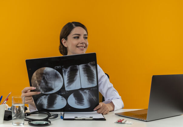 壁板面带微笑的年轻女医生穿着医用长袍和听诊器坐在办公桌旁 拿着医疗工具和笔记本电脑 一边看一边拿着x光片笔记本电脑微笑抱着