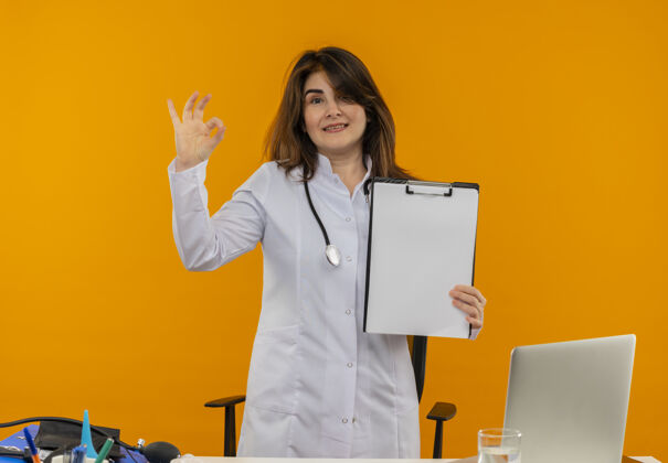 微笑微笑的中年女医生穿着医用长袍和听诊器站在办公桌后面 手里拿着医疗工具和笔记本电脑 手里拿着剪贴板 做着“ok”的手势剪贴板中年人笔记本电脑