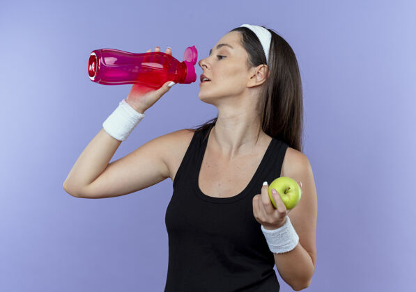 喝酒戴着头巾的年轻健身女士 在蓝色背景下站着 锻炼后拿着绿色苹果饮用水抱着站着锻炼