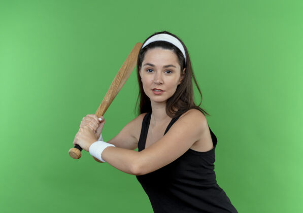 挥杆戴着头巾 挥舞着棒球棒 自信地站在绿色背景下的年轻健身女士健身棒球头带