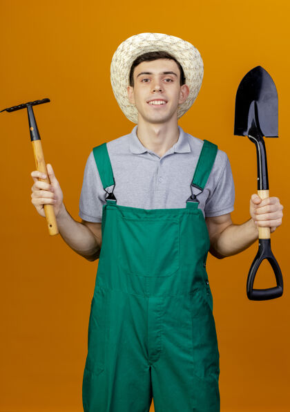 帽子微笑着的年轻男园丁戴着园艺帽 拿着耙子和铲子 在橙色的背景上与复制空间隔离开来年轻微笑园艺