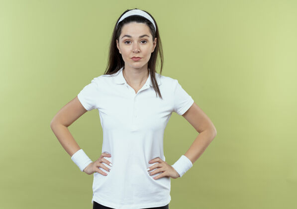 手臂戴着头巾的年轻健身女士站在灯光背景下 用自信的表情看着相机 双臂放在臀部年轻站立头带