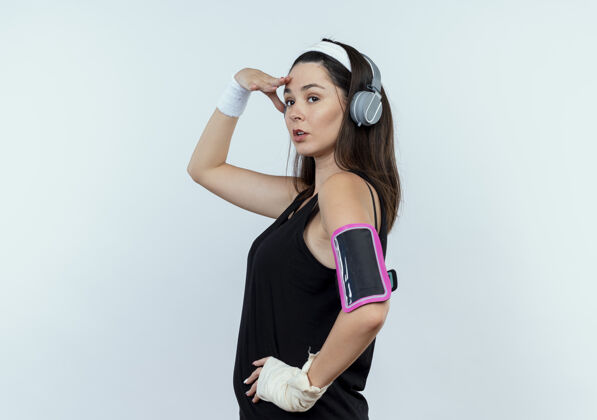 头戴着头戴耳机和智能手机袖标的年轻健身女手戴着头站在白色背景上遥望远方客场臂带头带