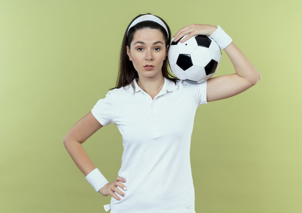 相机戴着头巾 肩上扛着足球的年轻健身女士站在灯光背景下 带着自信的表情看着摄像机头带肩膀表情