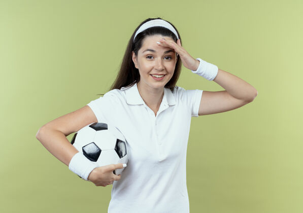 立场戴着头巾拿着足球的年轻健身女士站在灯光背景下 看起来很困惑 因为她站错了健身头年轻