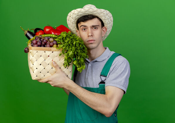 空间自信的年轻男园丁戴着园艺帽 拿着蔬菜篮子 在绿色的背景上与复制空间隔离开来持有帽子篮子
