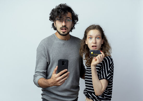 情侣一对年轻漂亮的情侣看着镜头困惑的男人拿着智能手机站在他的女朋友旁边谁拿着信用卡在白色的背景手机年轻困惑
