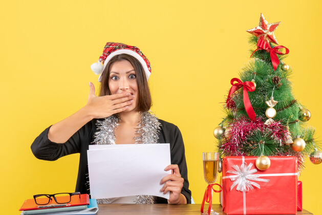圣诞老人穿着西装 戴着圣诞老人帽 戴着新年装饰品的商务女士看着一些令人惊讶的东西 坐在办公室的一张桌子旁 桌上放着圣诞树树惊喜漂亮