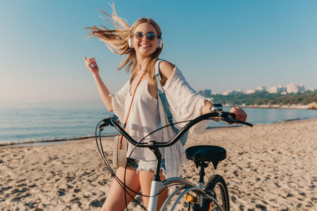 积极年轻迷人的金发微笑的女人 戴着耳机骑着自行车走在沙滩上听音乐放松音乐休闲