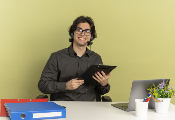 眼镜戴着耳机 戴着眼镜 面带微笑的年轻上班族坐在办公桌旁 手里拿着笔记本电脑 手里拿着剪贴板 看着隔离在绿色背景上的相机 还有复印空间微笑剪贴板笔记本电脑
