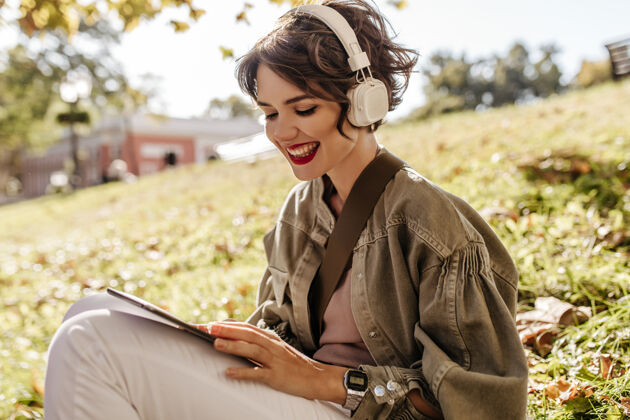 黑发穿着夹克戴着耳机坐在草地上的可爱女人快乐的卷发女人拿着智能手机站在外面身体公园成人