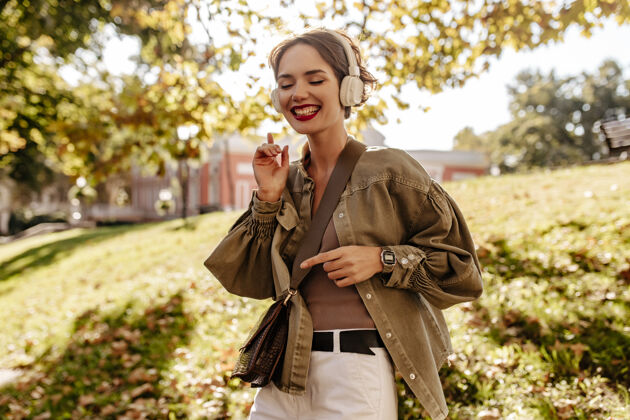 时尚快乐的女人穿着橄榄色夹克和白色牛仔裤在外面微笑波浪形头发的女人戴着耳机和手提包在户外听音乐城市走道肖像
