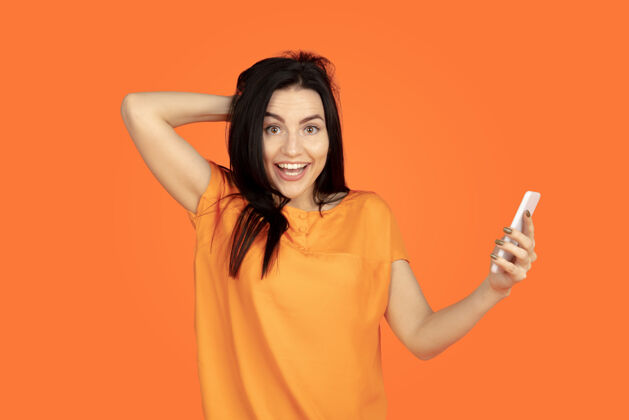 女性橙色工作室背景上的白种人年轻女子肖像穿着衬衫的漂亮深色女性模特人类情感的概念 面部表情 销售 广告广告空间自拍 赌赢员工手势公司