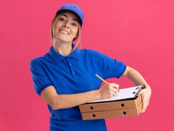 递送穿着制服的年轻漂亮的女送货员微笑着拿着比萨饼盒 用粉色墙上的钢笔在剪贴板上写字人书写漂亮