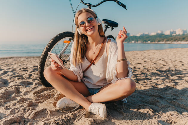 活跃时尚年轻迷人的金发微笑的女人坐在沙滩上骑着自行车戴着耳机听音乐休闲服装海岸