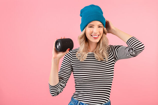 现代年轻漂亮的女人拿着无线扬声器听音乐穿着条纹衬衫和蓝色帽子微笑快乐积极的心情摆在粉红色的墙上享受装置微笑