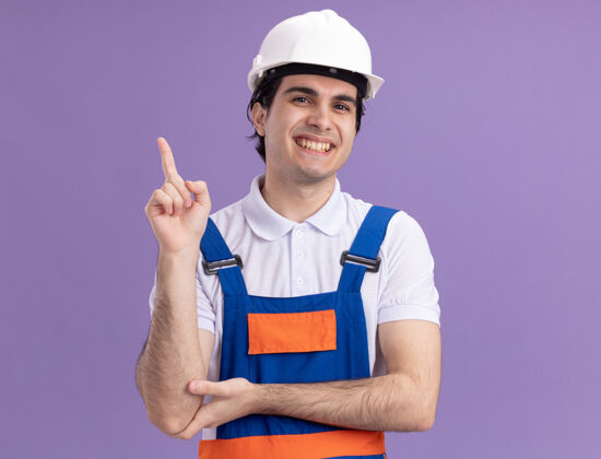 站立年轻的建筑工人穿着建筑制服 戴着安全帽 微笑着看着前面 用食指指着站在紫色墙上的一边指点手指目录