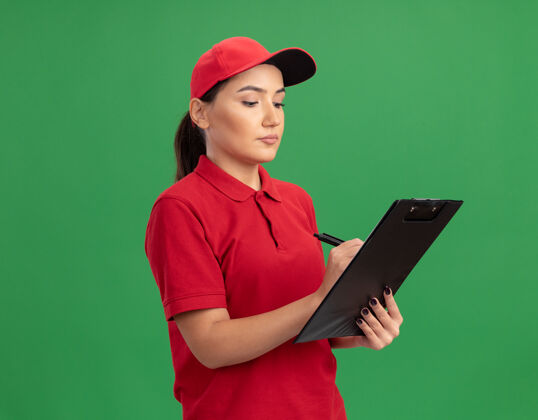 立场身穿红色制服 头戴便帽的年轻女送货员站在绿色的墙上 用剪贴板写着一张严肃的脸写作严重交货