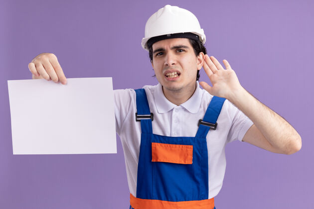头盔年轻的建筑工人穿着建筑制服 戴着安全帽 拿着一张空白的纸 用手捂着耳朵看着前面 试图听站在紫色墙上的流言蜚语建筑工人耳朵空白