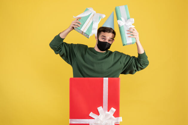 礼物正面图戴着派对帽的年轻人拿着礼物站在黄色背景的大礼盒后面前面购物礼品盒