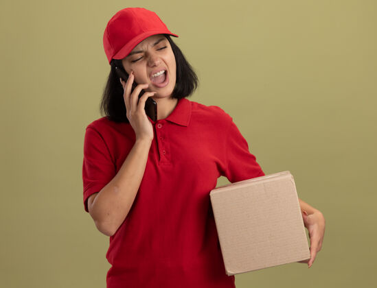 手机身穿红色制服 头戴鸭舌帽的年轻送货员站在轻质墙上一边拿着纸板箱一边喊着手机拿着制服站着