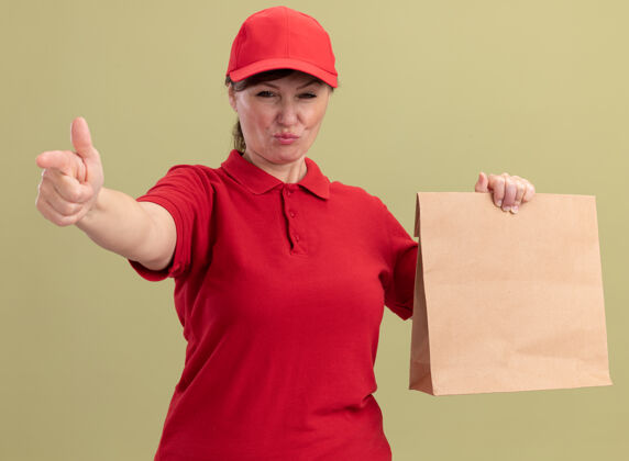 立场身穿红色制服 戴着礼帽的中年送货妇女站在绿色的墙上 用食指指着前面 表情严肃自信地看着前面女人包装严重