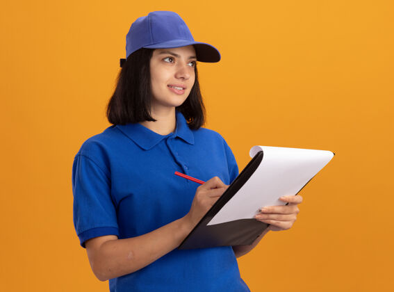 持有身穿蓝色制服 戴着帽子的年轻送货女孩站在橙色的墙上 手里拿着空白页和铅笔的剪贴板 面带微笑地看着一旁旁边立场帽子