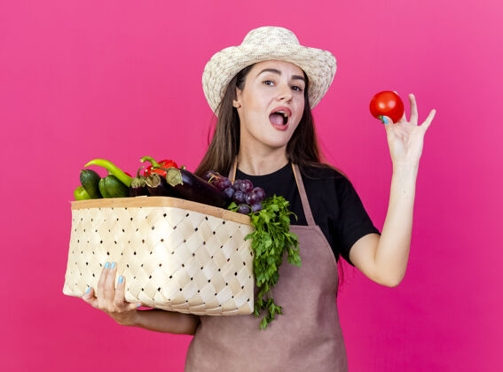 举行一个穿着制服的漂亮的园丁女孩戴着园艺帽 手里拿着一个蔬菜篮子 篮子里的西红柿被隔离在粉红色的背景上番茄花园蔬菜