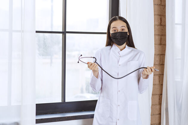 设备戴着黑色口罩的医生拿着听诊器站在窗边 看上去很困惑惊险工具妇女
