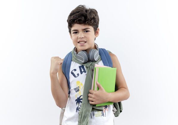 小学生看着镜头 一个背着书包 戴着耳机 手拿书本 手指交叉的小男孩被隔离在白色背景上小包手指