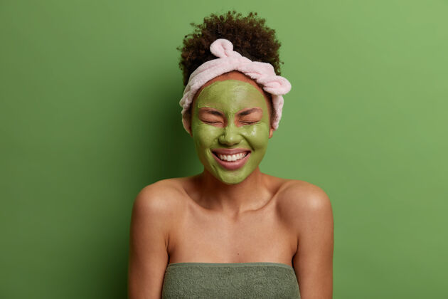 高兴年轻的喜出望外的女人用天然粘土面膜来减少粉刺 笑容灿烂 拥有完美的白色牙齿 梳过卷发 戴着头带 露出赤裸的肩膀 隔离在绿色的墙上女士毛巾完美