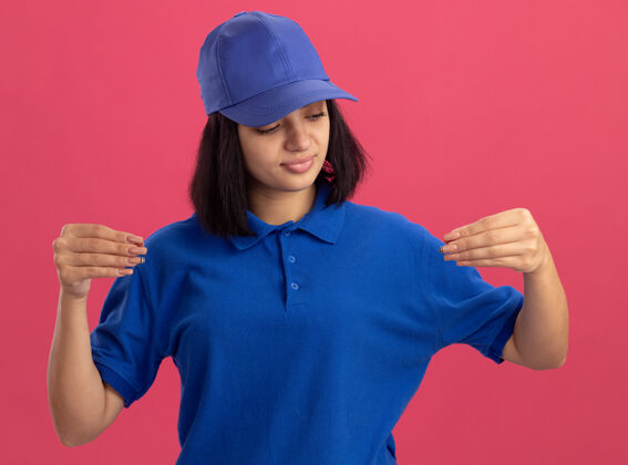 手指身穿蓝色制服 戴着帽子的年轻送货女孩自信地用手指做手势 站在粉色墙壁上的肢体语言概念交货制服年轻