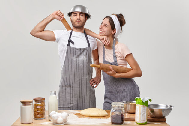 配料骄傲的男人和他快乐的妻子在厨房忙碌着 穿着围裙 完成面团的制作 一起烤面包 使用秘方 靠着桌子旁边的白墙站着新鲜的产品糕点产品准备