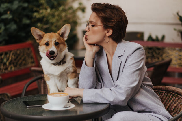 女性短发女士在咖啡馆喝咖啡 看着她的狗穿着灰色夹克的迷人女士在外面和柯基犬一起休息座位狗女人
