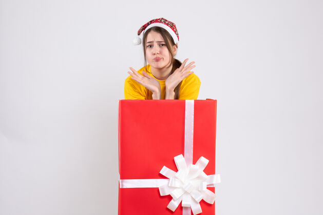 人前视图困惑可爱的女孩与圣诞老人帽子站在大礼物后面购物帽子圣诞老人