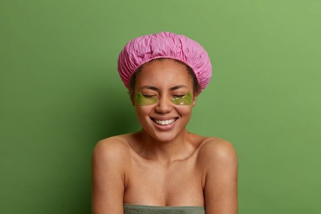 过程快乐的美国黑人妇女戴着浴帽 裹着毛巾 淋浴后敷在眼罩下 关心皮肤 对着绿色的墙壁摆姿势宠爱身体外观