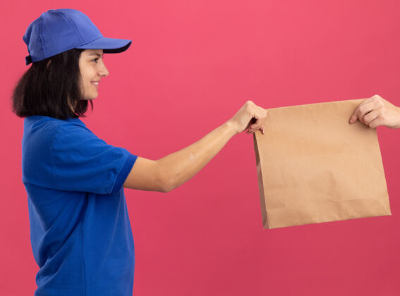年轻身穿蓝色制服 头戴帽子的年轻送货员站在粉红色的墙上 微笑着向顾客赠送纸质包装包装顾客女孩