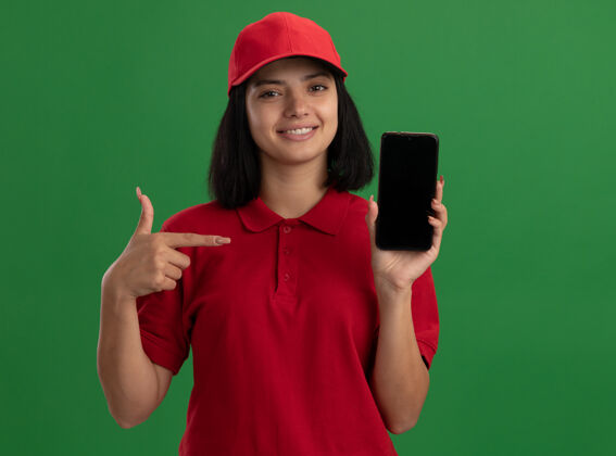 目录身穿红色制服 戴着帽子的年轻送货女孩站在绿色的墙上 用食指指着智能手机 自信地微笑着指着手机站着