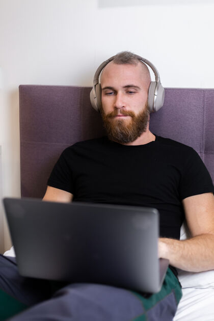 胡须一个留着大胡子的白种人戴着耳机在卧室的床上工作 在家里用笔记本电脑 打字 思考卧室积极手机