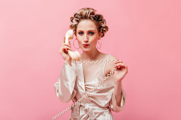 情感在粉红色的墙上 一个带着卷发的调情女人咬着嘴唇 对着固定电话说话时尚衣服杯子
