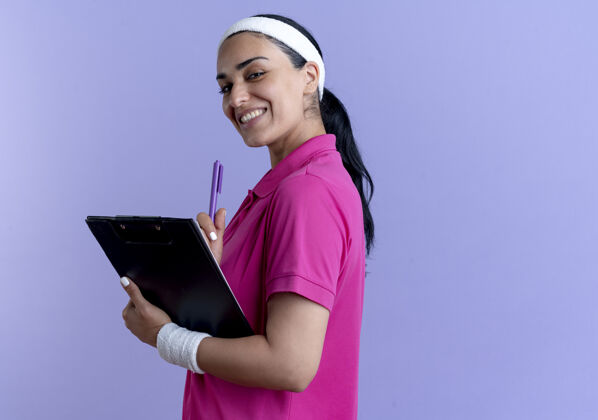 钢笔年轻微笑的白人运动女性戴着头带和腕带站在一边拿着笔和剪贴板在紫色的复制空间年轻腕带空间