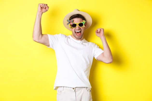 工作室旅游和生活方式的概念快乐幸运的家伙赢得旅行 高兴和穿着节日服装 夏季帽子和太阳镜 黄色背景黄色帅气男人