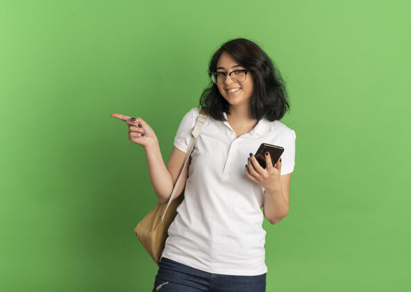 点年轻的高加索女学生 面带微笑 戴着眼镜 背着书包 站在一旁 手里拿着手机 放在绿色的空间里空间绿色包