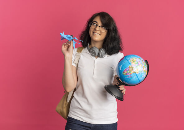 耳机年轻可爱的白人女学生戴着耳机戴着眼镜背着包拿着飞机和地球仪仰望着粉色的空间包请漂亮
